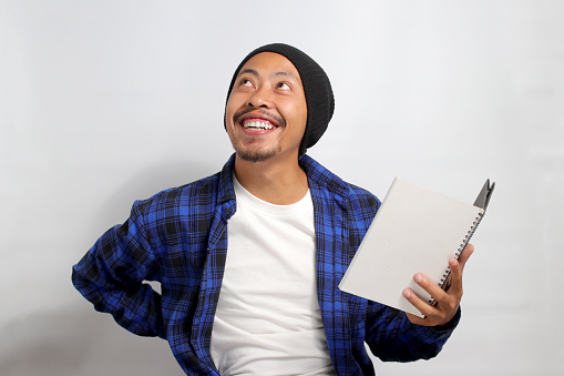 An enthusiastic young Asian student, dressed in a beanie hat and casual shirt, holds a book while looking at an empty space meant for copy or text, seemingly lost in imagination or contemplation