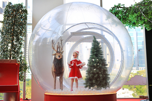 Adorable little child girl in santa costume dress playing in big winter Snow globe with reindeer on christmas time. Merry Xmas and happy new year.