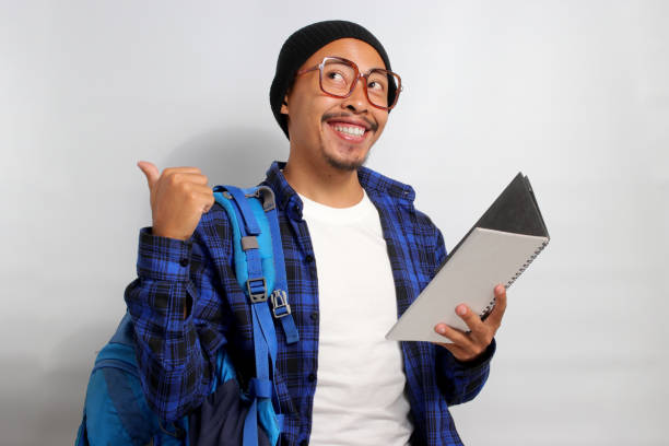 istock Excited young Asian student, wearing eyeglasses and a backpack, is pointing at an empty copy space 1861369248