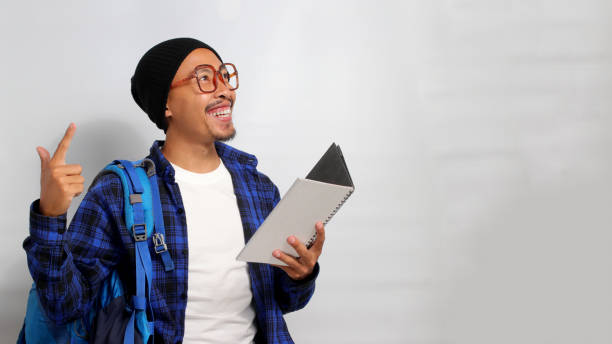 istock Excited young Asian student, wearing eyeglasses and a backpack, is pointing at an empty copy space 1861374018
