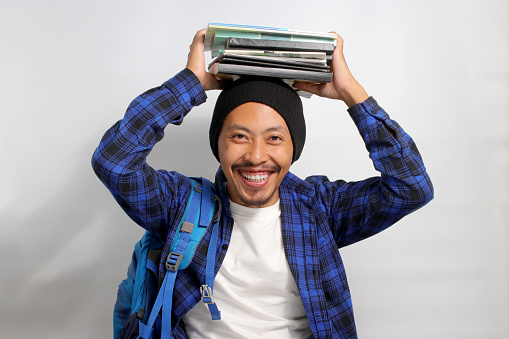 A cheerful young Asian male student, dressed in a beanie hat and casual shirt, carrying a backpack, is holding a stack of books on his head while smiling and looking at the camera, isolated on white