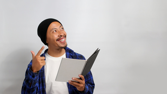 An excited young Asian student, dressed in a beanie hat and casual clothes, is smiling while pointing aside at an empty copy space, standing against a white background