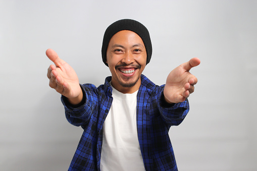 Friendly, smiling young Asian man extends his arms for a hug towards the camera, gesturing a COME HERE sign, inviting an embrace with a toothy smile while standing against white background