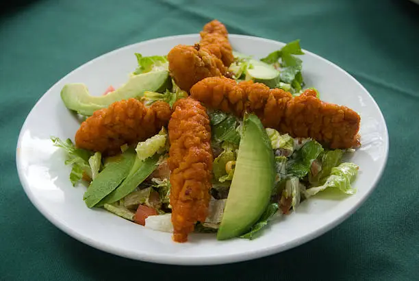 Spicy fried chicken upon a salad of greens and avocado in a white dish on a green tablecloth.