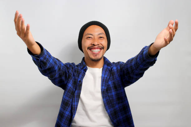Overjoyed Asian man stretches his arms wide open towards the camera Overjoyed Asian man, dressed in a beanie hat and casual shirt, stretches his arms wide open towards the camera, offering a hug gesture, inviting an embrace while standing against a white background male is standing and shows outstretched hand with open palm stock pictures, royalty-free photos & images