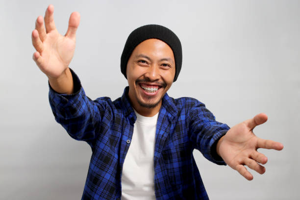 Overjoyed Asian man stretches his arms wide open towards the camera Overjoyed Asian man, dressed in a beanie hat and casual shirt, stretches his arms wide open towards the camera, offering a hug gesture, inviting an embrace while standing against a white background male is standing and shows outstretched hand with open palm stock pictures, royalty-free photos & images