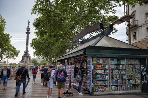 News stand and gift shop kiosk on La Rambla street, Barcelona, Spain.  Tourists exploring city and buying souvenirs, Christopher Columbus Monument in the background.