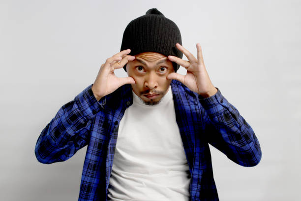 Sleepy, tired young Asian man in a beanie hat and casual outfit is struggling to open his eyes, displaying signs of morning fatigue or exhaustion, isolated on white background.