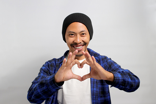 Attractive Asian man, dressed in a beanie hat and casual shirt, exudes happiness as he creates a romantic heart gesture with his hand, expressing tender feelings, standing against white background