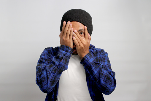 A surprised young Asian man, dressed in a beanie hat and casual shirt, is covering his face and eyes with his hand, peeking through his fingers in a shocked and embarrassed expression