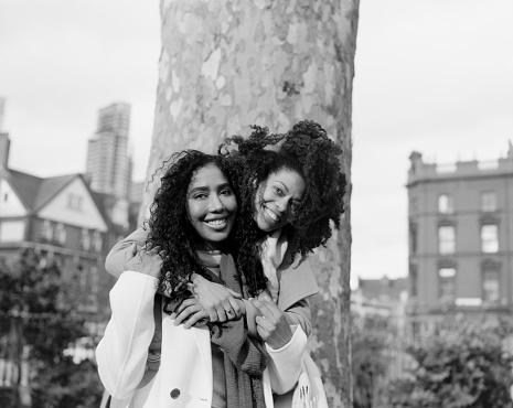 Analog portrait of two female friends hugging each other in front of big tree in a street of London. One is hugging the other one from behind and both of them are smiling happily.