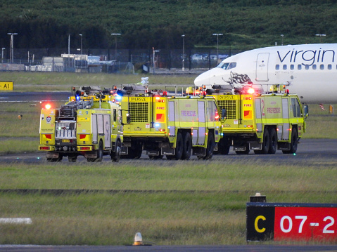 The fire engines of Sydney Kingsford-Smith Airport rushed to a Virgin Australia B737-8FE plane, registration VH-VFL, after landing as flight VA877 from Melbourne.  They remained in place for five minutes while the plane remained stationary.  The plane taxied to the domestic terminal and the fire engines followed her.  The sign in red and white numbers refers to the east-west runway.  This image was taken from Shep's Mound, Ross Smith Avenue, Mascot on a hot evening at sunset on 14 December 2023.