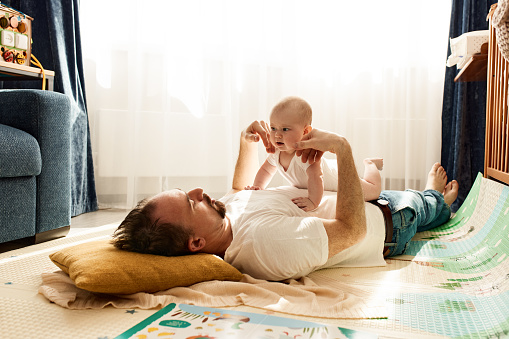A cute joyful kid and his father in a white T-shirt and jeans lie on the floor in a bright room and play together. Happy fatherhood and childhood.