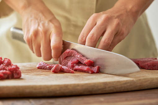 Crop professional female chef in process of cutting beef on wooden chopping board with sharp knife while cooking
