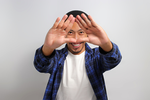 Young Asian man, dressed in a casual shirt and wearing a beanie hat, is creating a triangle shape with his hands, symbolizing the concept of environmental protection, recycling, and reusing