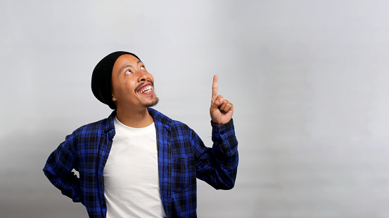 An excited young Asian man, dressed in a beanie hat and casual shirt, is pointing upward at an empty space while standing against a white background