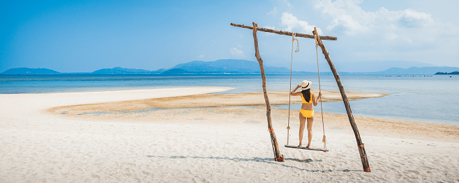 Summer travel vacation concept, Happy traveler asian woman with bikini and hat relax in swing on beach in Koh Rap, Samui, Surat Thani, Thailand