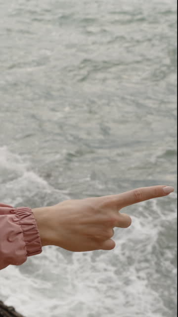 Vertical video. A Woman Stands Among the Rocks by the Sea in Strong Wind, Wearing a Pink Hooded Raincoat, and Points Somewhere with Her Finger.