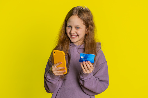 Portrait of happy teen child girl kid using credit bank card and smartphone while transferring spending money, purchases online shopping, ordering food delivery. Children isolated on yellow background