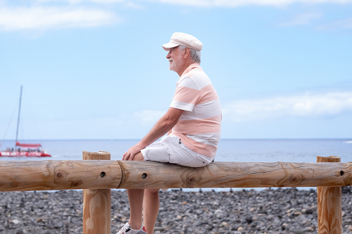 Portrait of relaxed caucasian bearded man with hat sitting at the beach looking at the horizon while enjoying vacation or retirement. Horizon over the water, copy space