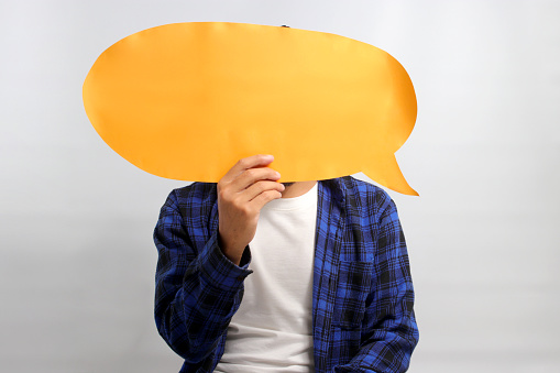 Asian man, concealing his face behind a speech bubble held in front of him, stands against a white background, depicting the concepts of information, thoughts, quotes, opinions, reviews, and feedback