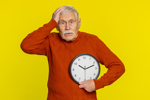Senior old man with anxiety checking time on clock, running late to work, being in delay, deadline. Elderly grandfather looking at hour, minutes, worrying to be punctual isolated on yellow background