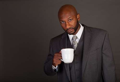 Young black business man on a grey background wearing a shirt tie and vest looking at the camera with coffee cup