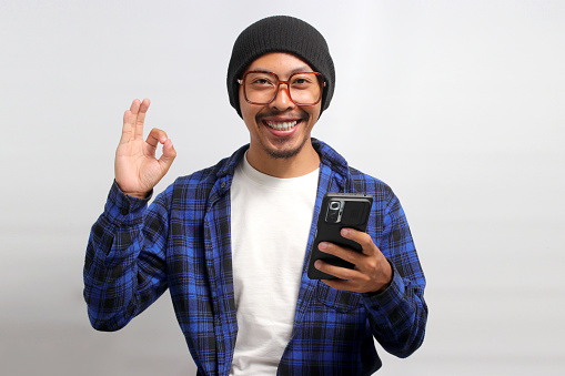 An Asian man, donning a beanie hat, casual shirt, and eyeglasses, flashes an OK sign towards the camera while holding a phone, conveying satisfaction, approval, and a positive review, white background