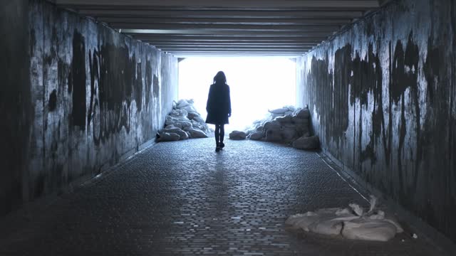 Silhouette woman walks along an underground passage turned into a bomb shelter towards the light. Rear view, shabby tunnel, sandbags. War in Ukraine.