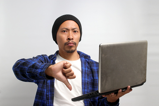 A displeased young Asian man, dressed in a beanie hat and casual shirt, expresses dissatisfaction with a thumbs down gesture while holding a laptop in his hand, isolated on a white background