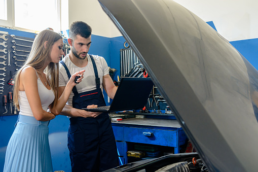 A Car Mechanic is Working on the Car Engine in a Car Service and Trying to Explain a Failure to a Young Female Customer and Holding a Laptop.