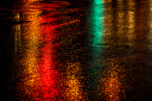 Abstract, wet asphalt road colorfully illuminated at night by red car taillights and traffic signal lights.