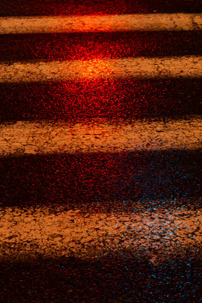 Abstract, textured, cracked section of an asphalt, black crosswalk on a rainy night illuminated by colorful, artificial light.