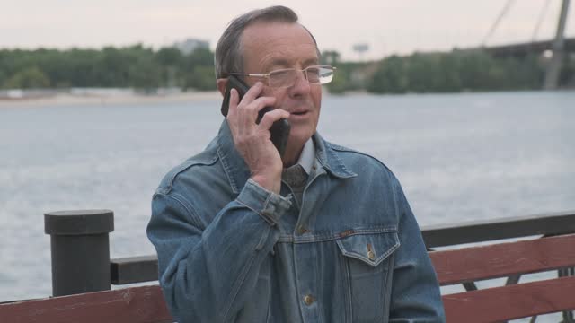 Elderly man answers the phone. Old people using modern smartphones. Front view, close-up, outdoor, daytime, overcast.