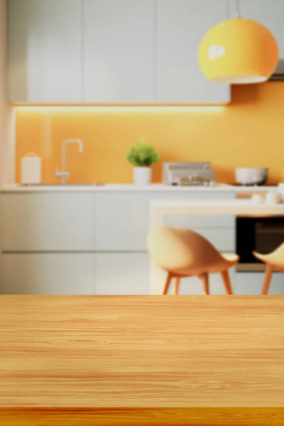 Defocused modern kitchen with empty wooden table or countertop in foreground for object placement.  Cooking pans and food preparation in background.  Stainless steel appliances. No people.