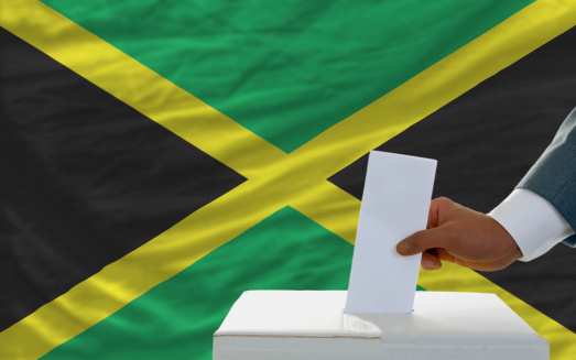 man putting ballot in a box during elections in jamaica in fornt of flag
