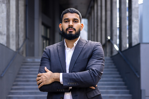 Portrait of a young Muslim male lawyer standing in a business suit outside a courthouse and looking confidently into the camera with his arms crossed.