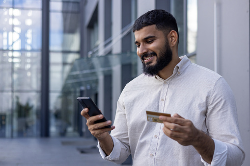 Smiling young male businessman standing near office in white shirt, using phone and credit card.
