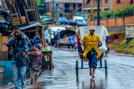 Ambositra town, Madagascar, October 17, 2017: A rickshaw puller is seen pulling a Pulled rickshaw (pousse-pousse) in the rain with the riders covered with a plastic sheet for protection from the rain