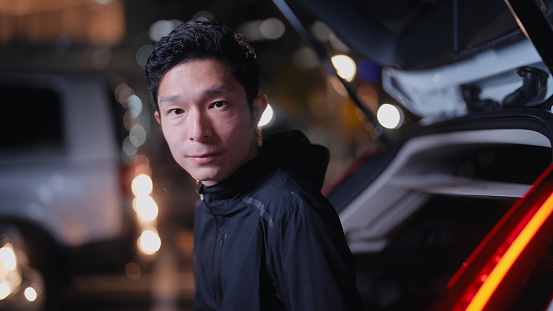 A portrait of a male  Asian athlete sitting on his car trunk and getting ready for training and running in a parking lot in the city at night.