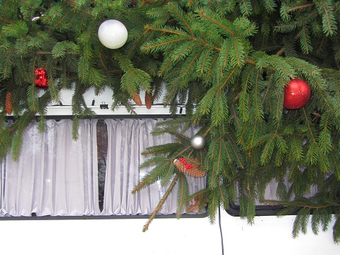 Christmas tree branches are decorated with New Year's toys. The cones are hanging. All this decoration lies on the roof of the car. White car curtains cover the elongated windows of a white car