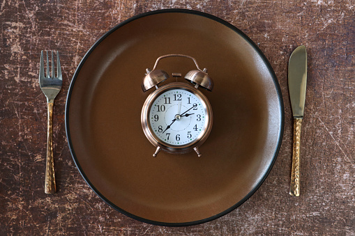 Stock photo showing close-up, elevated view of a healthy eating and intermittent fasting diet concept depicted by a plate containing a double bell alarm clock.