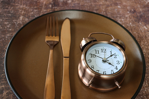 Stock photo showing close-up, elevated view of a healthy eating and intermittent fasting diet concept depicted by a plate containing a double bell alarm clock and stainless steel knife and fork.
