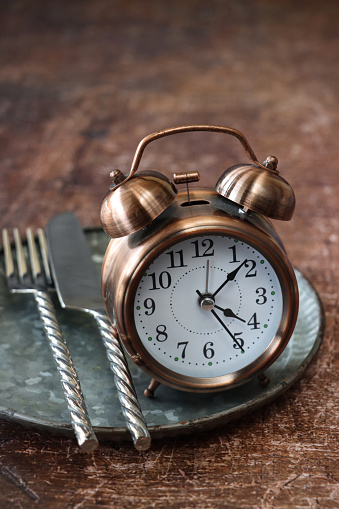 Stock photo showing close-up view of a healthy eating and intermittent fasting diet concept depicted by a plate containing a double bell alarm clock and stainless steel cutlery.