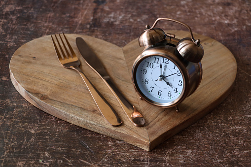 Stock photo showing close-up view of a healthy eating and intermittent fasting diet concept depicted by a wooden heart-shaped board containing a double bell alarm clock and stainless steel knife and fork.