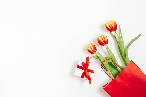 Flat lay composition with red yellow tulips in a red paper bag with gift on a white background with copy space. Valentin Day, Birthday, Mother day