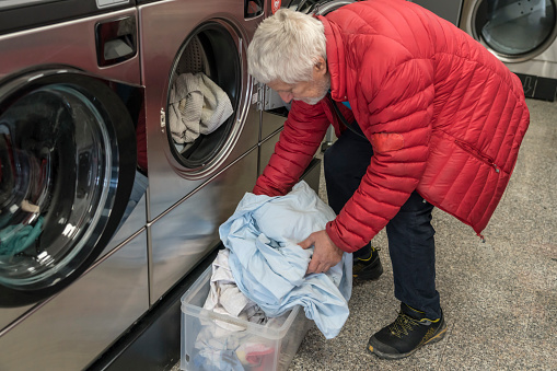 Senior men putting laundry into the washing machine at  public laundromat service.
