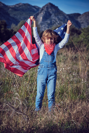 Cute preteen boy in denim overall standing in grassy meadow with waving American national flag and looking at camera