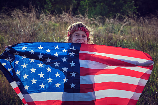 Black-haired girl covered with a large American flag, in the garden of her house. Concept of celebration, independence day, United States of America, 4th of July, patriotism and American pride.