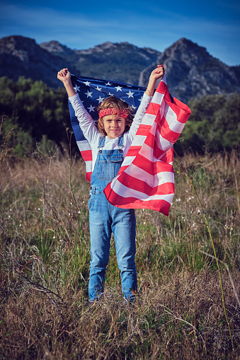 Cheerful preteen boy standing in grassy meadow with raised hands and flag of United States of America and looking at camera